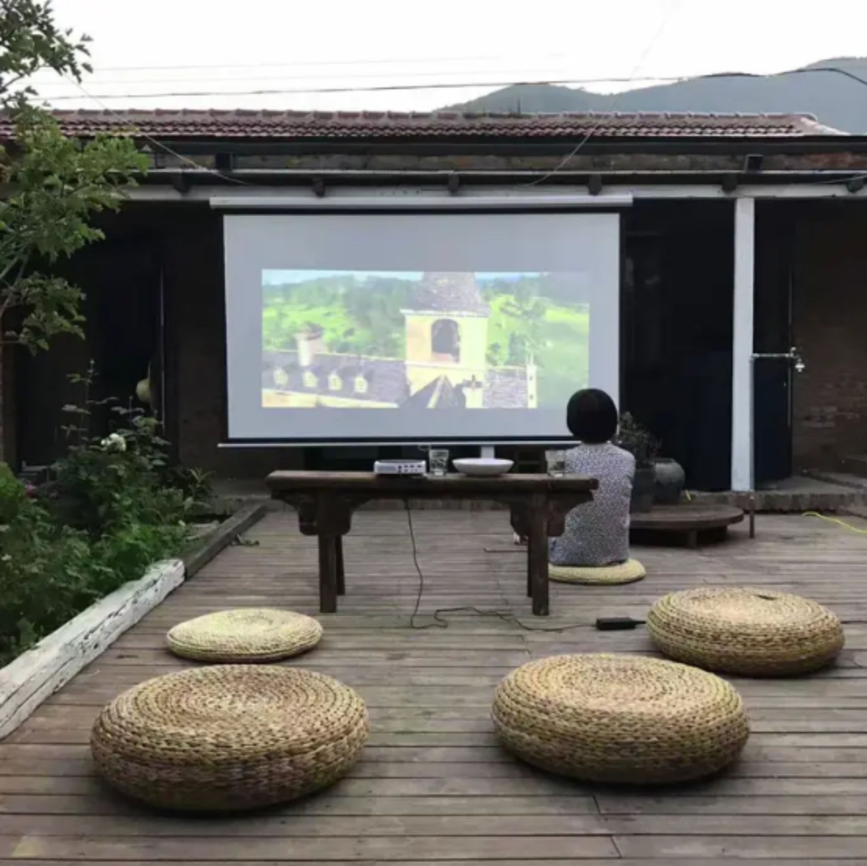 Una persona sentada en un taburete tejido frente a una pantalla de proyector que muestra un paisaje.