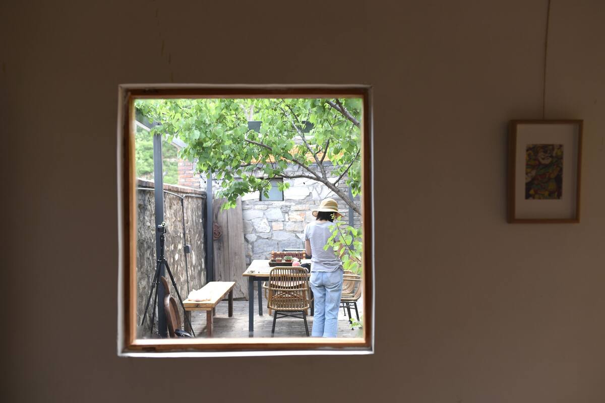 Habitación con ventana que da a un patio con mesa y sillas, un trípode y un muro de piedra.