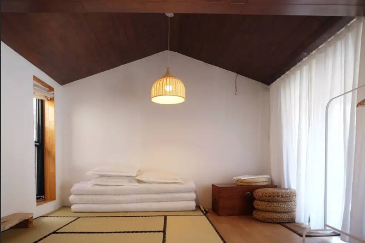 Una habitación con techo inclinado, lámpara colgante, una pila de toallas blancas, una mesita de madera y una ventana con cortinas.