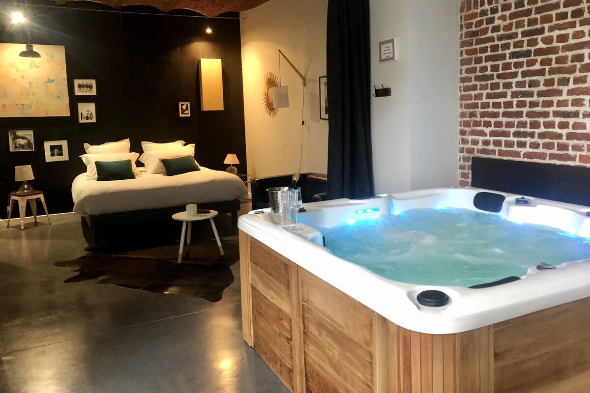 A modern hotel room with a large bed, a wooden hot tub, and a brick wall.