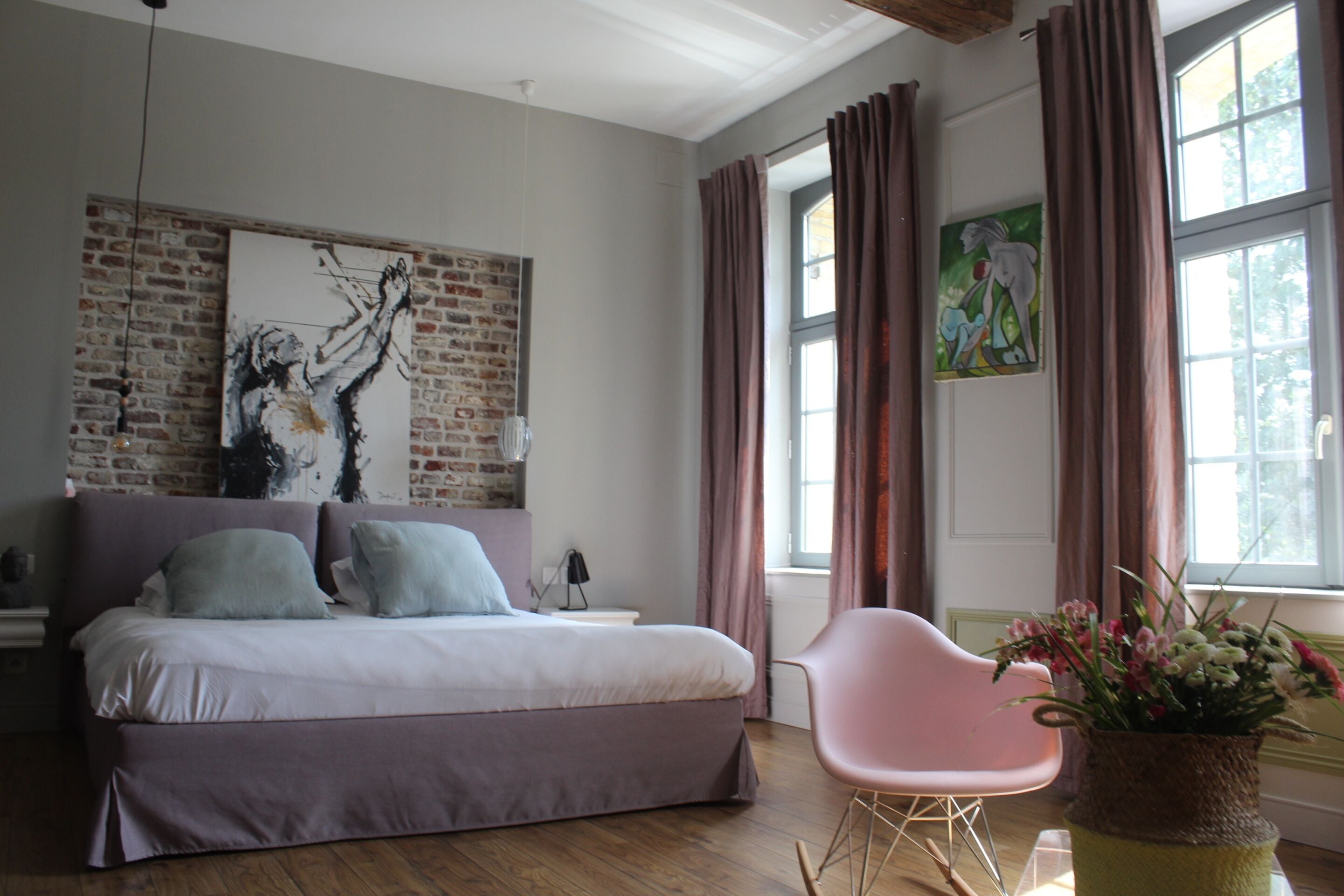 A bedroom with a bed, a chair, a window with curtains, and artwork on the walls.