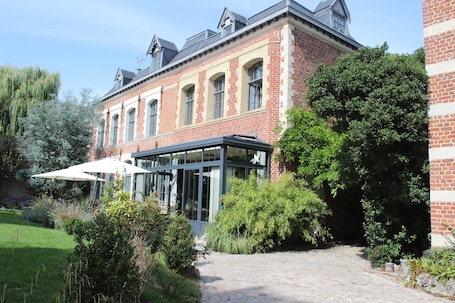 A brick building with a glass conservatory, a paved pathway, and lush greenery.. Maison Mathilde