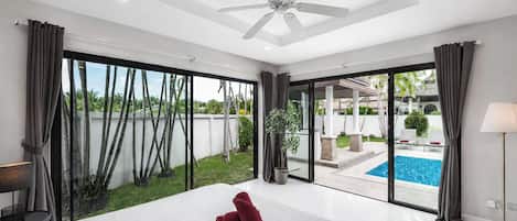 Une chambre spacieuse avec un grand lit, un ventilateur de plafond et une vue sur une piscine, visible à travers de grandes portes coulissantes.