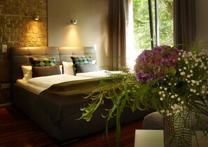 A modern hotel room with a large bed, a brick accent wall, and a view of greenery outside. - Trafohaus am Kaiserbahnhof (Bruehl)