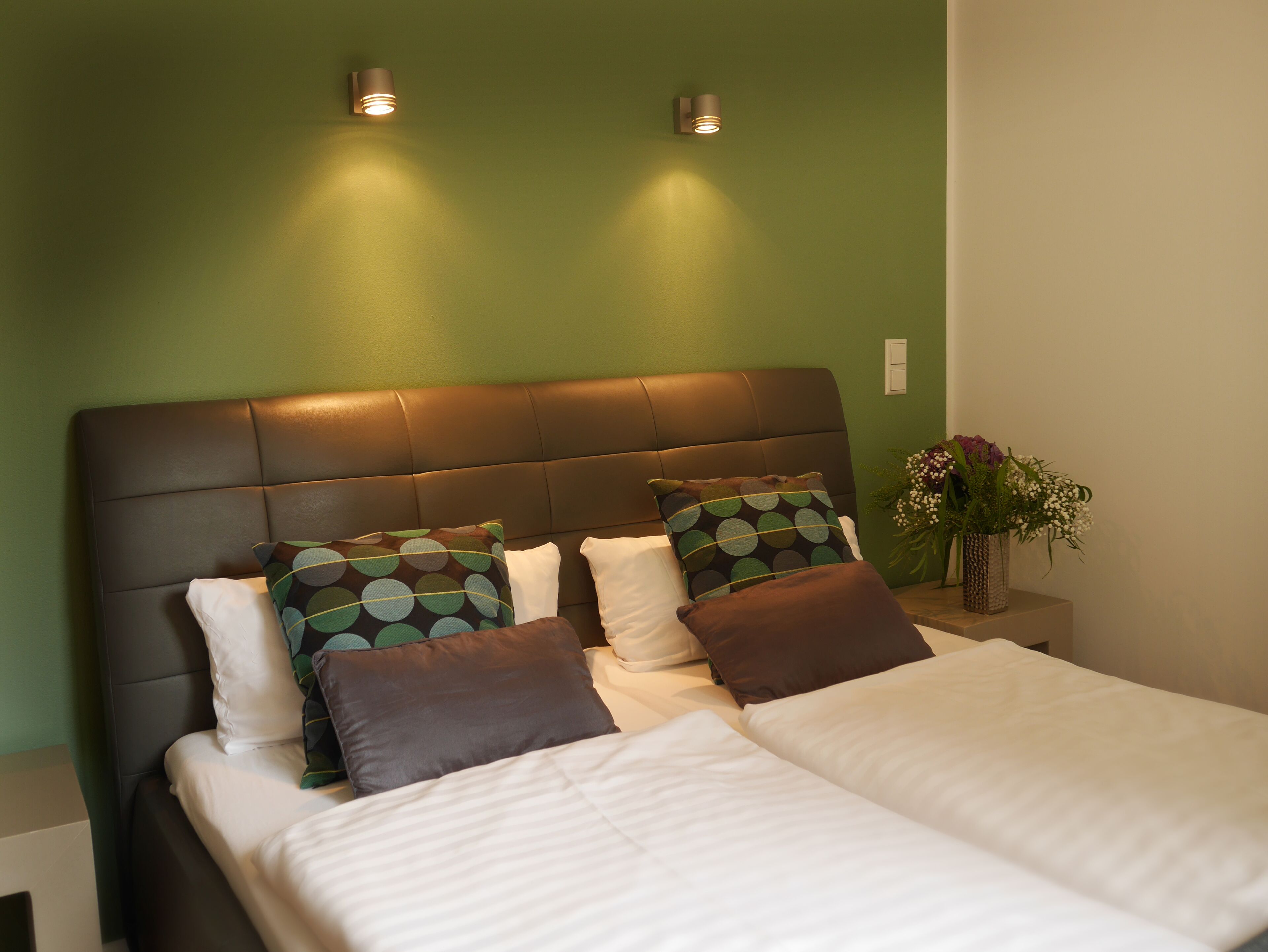 Cama bem arrumada com roupa de cama branca e marrom, cabeceira escura e duas luminárias fixadas na parede.