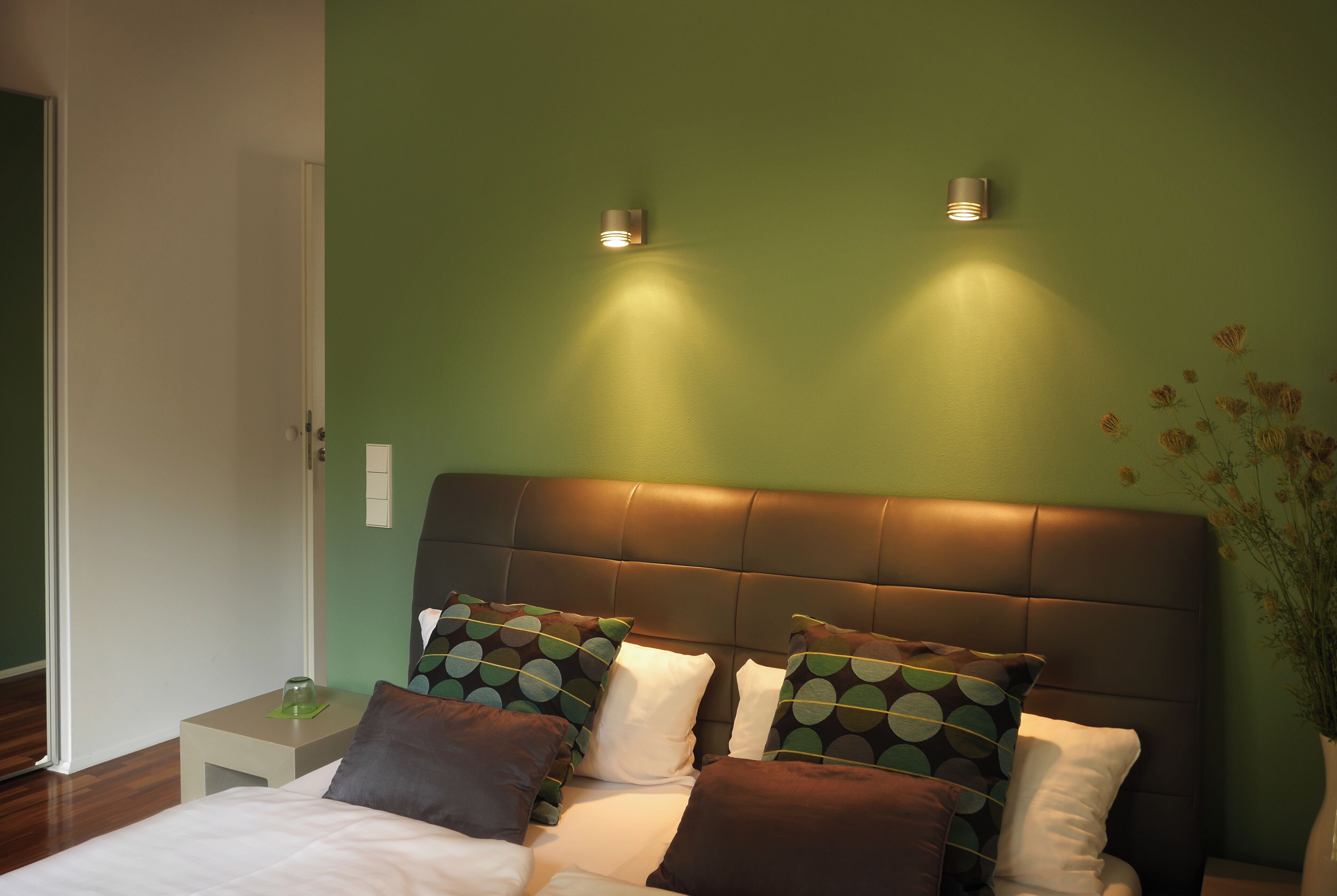 A neatly made bed with multiple pillows, a bedside table with a lamp, and a wall-mounted light fixture.