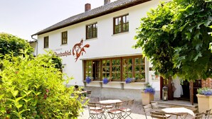 Un bâtiment à la façade blanche, une enseigne indiquant « Pension Thiembachthal », et des tables et des chaises disposées à l’extérieur pour les clients.