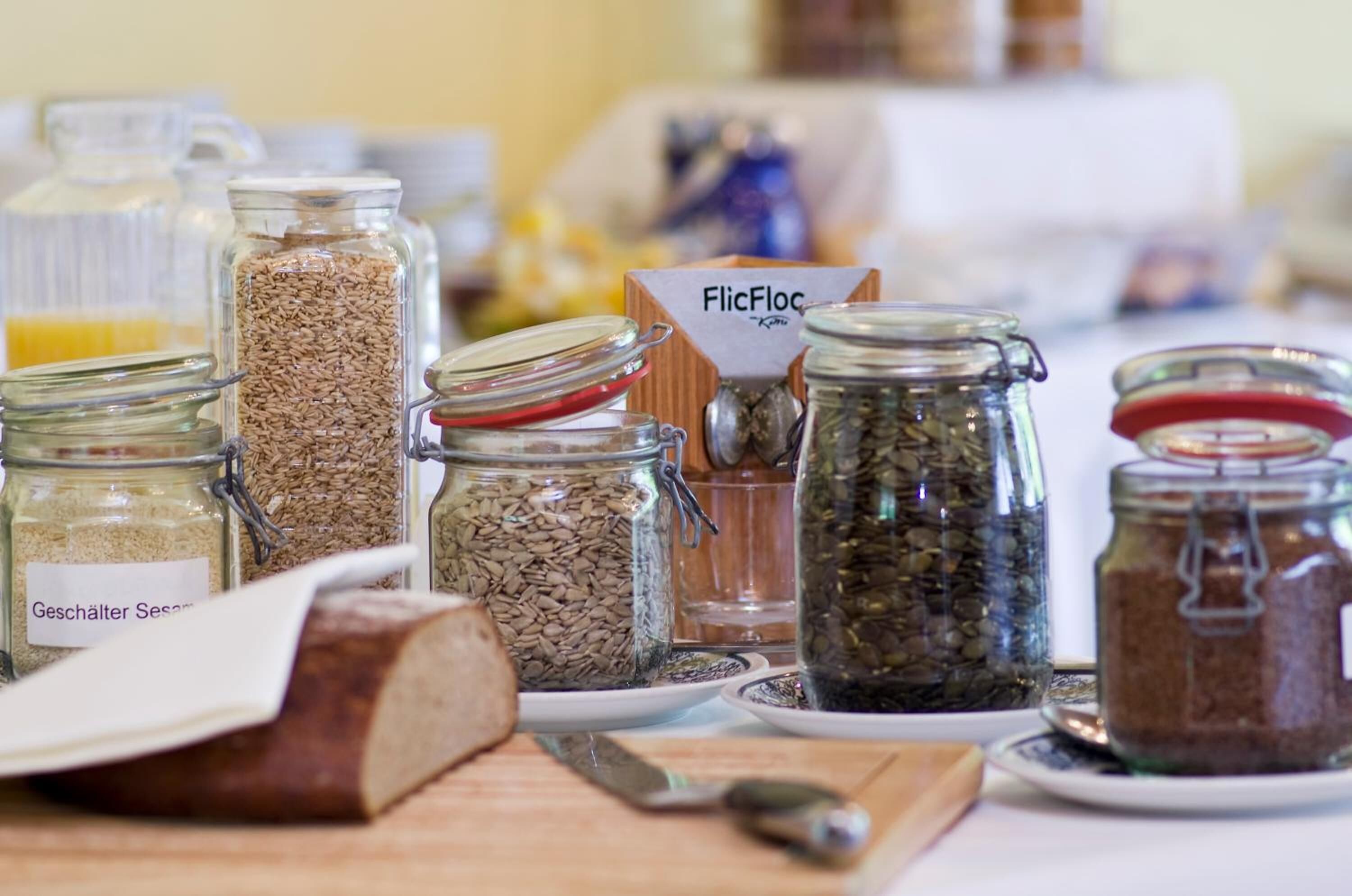 A variety of glass jars containing different grains and seeds, a loaf of bread, and a knife on a table.
