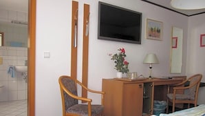 Habitación de hotel con cama, escritorio, silla, televisor, espejo y planta.