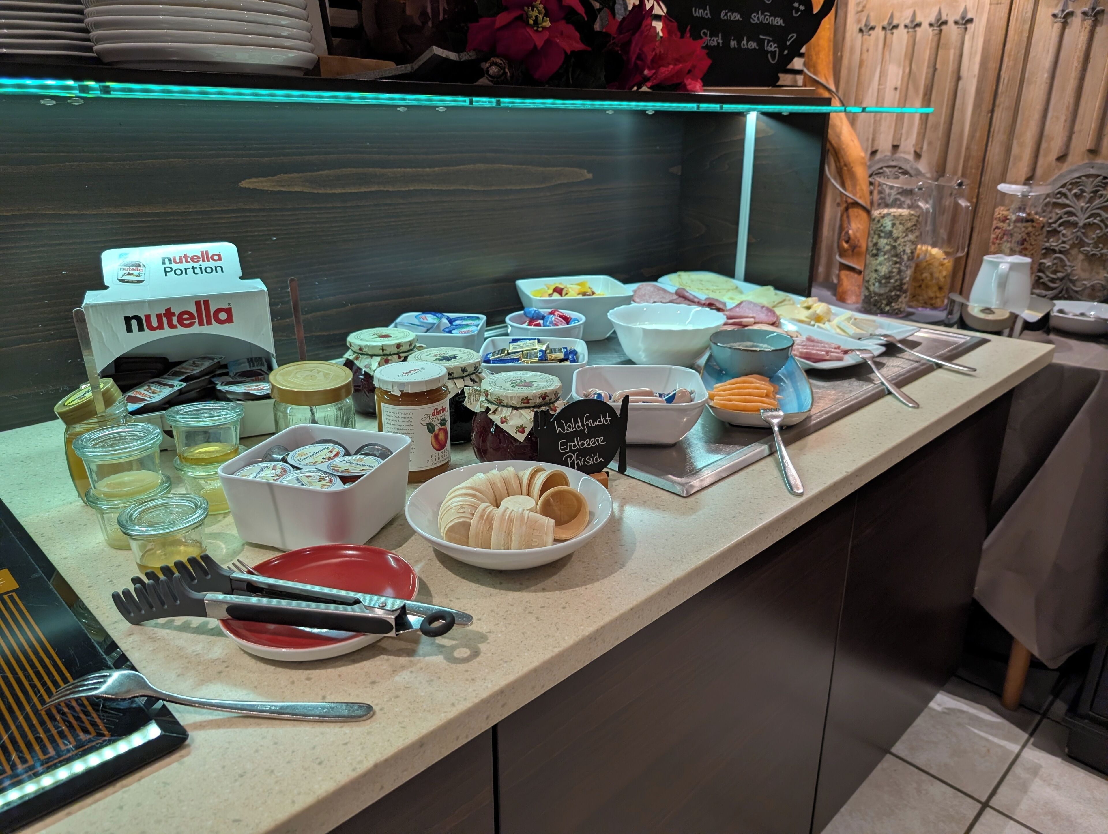 Ein Buffet mit verschiedenen Speisen, darunter Gläser mit Marmelade, eine Packung Nutella und Besteck.