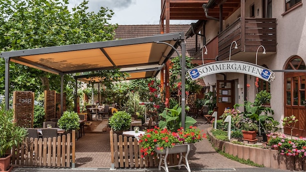 A covered outdoor seating area with tables and chairs, surrounded by potted plants and flowers. - Hotel-Gasthof Weißes Roß (Kleinostheim)
