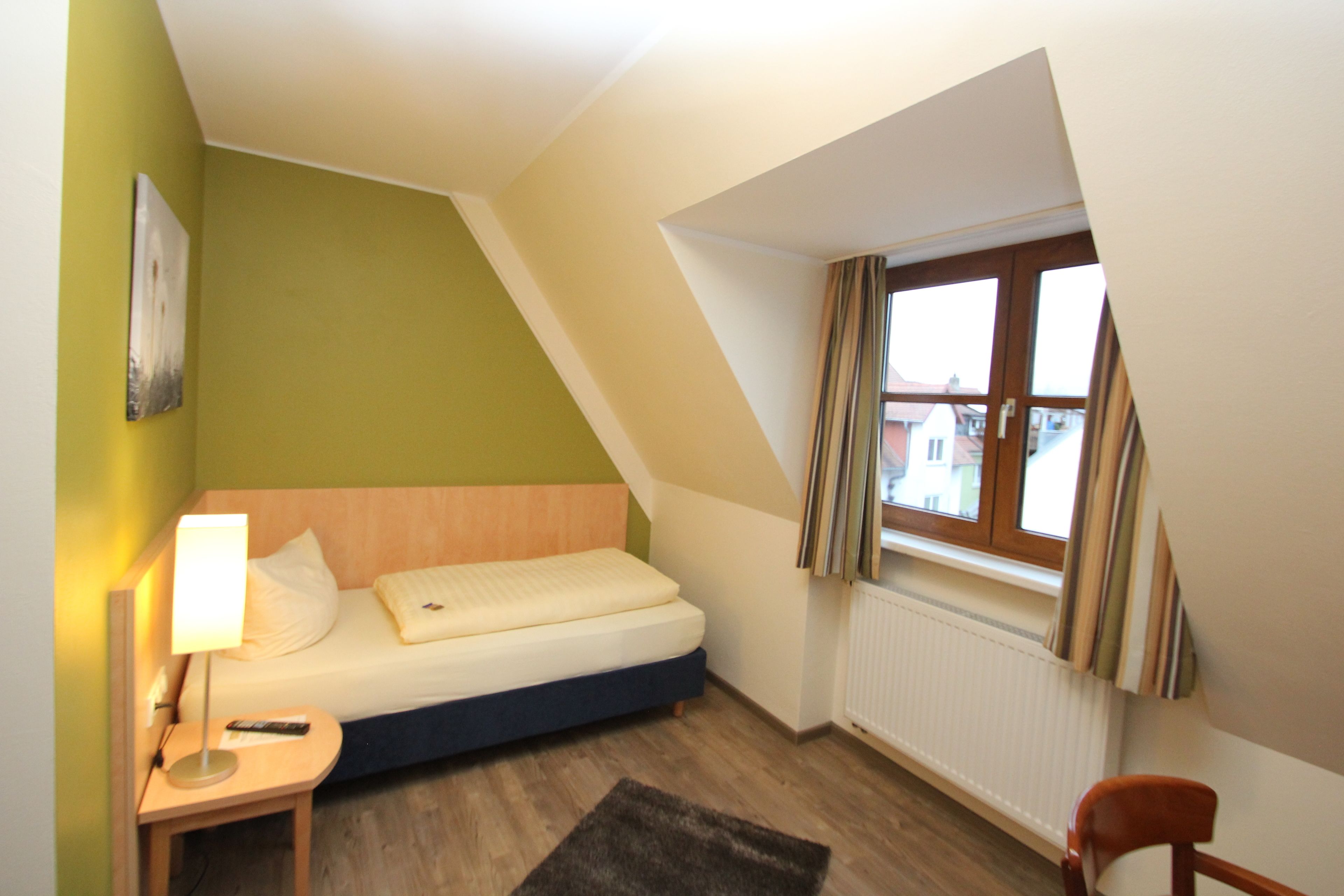 Una habitación pequeña y triangular con una cama, una mesita de noche, una silla y una ventana con cortinas.