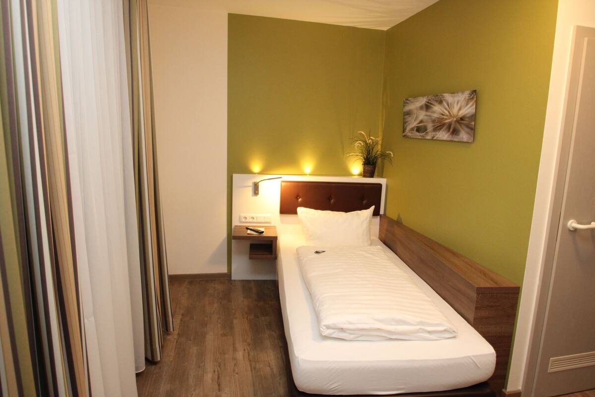 A small, neatly made bed with a wooden headboard, a bedside table with a lamp, and a wall-mounted artwork.