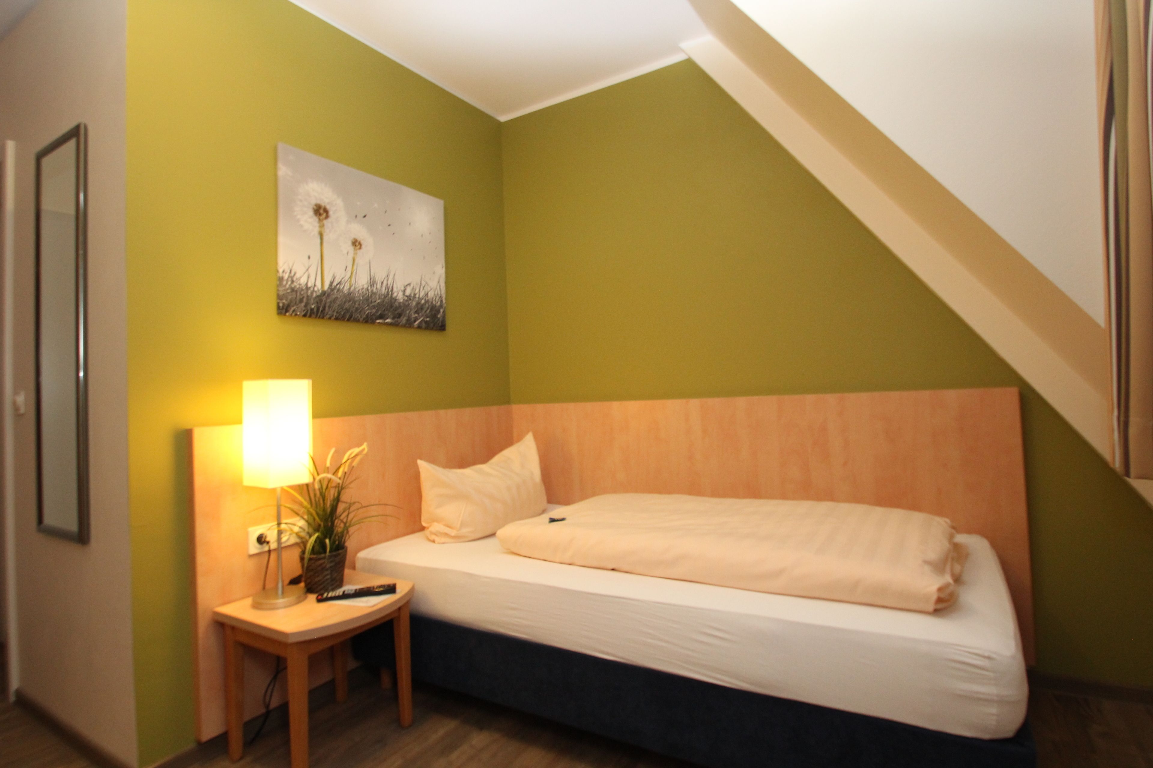 Habitación de hotel con cama, mesita de noche, lámpara, un cuadro en la pared y ventana con vista al cielo.