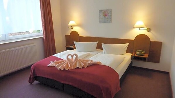 A hotel room with a bed, two bedside lamps, a window with curtains, and a radiator.