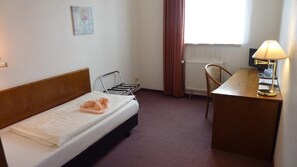 A hotel room with a single bed, a desk with a lamp, a chair, a radiator, and a window with curtains.