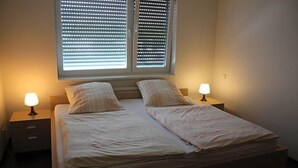 A double bed with two bedside tables, each with a lamp, in a room with a window with blinds. - Hotel Grand Tour (Wesseling)