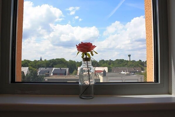 Vista despejada de un paisaje urbano a través de una ventana, con una sola rosa roja en un tarro sobre el alféizar.