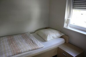 A small, single-bed room with a window, a bedside table, and a lamp.