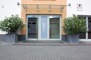 Entrada de hotel con puerta de vidrio, un cartel que dice 'GRAND TOUR' y dos plantas en macetas a ambos lados.