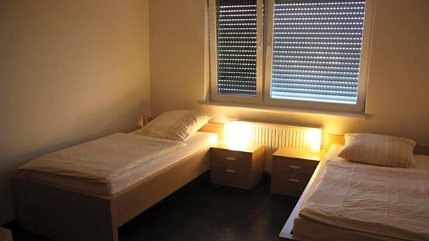 A bedroom with two single beds, a nightstand, and a window with blinds. - Hotel Grand Tour (Wesseling)
