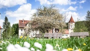 A building with a red roof, surrounded by a garden with white and yellow flowers, and a clear blue sky. - Hotel Schloss Blumenthal (Aichach)