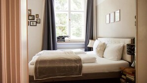 A bedroom with a bed, a window with curtains, a bedside lamp, and a wall with framed pictures. - Hotel Schloss Blumenthal (Aichach)