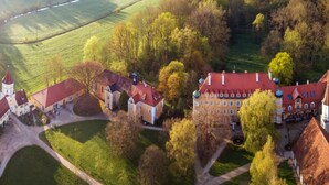 An aerial view of a historic estate with multiple buildings, including a large main house with a red roof, smaller structures, and a church with a tower. - Hotel Schloss Blumenthal (Aichach)