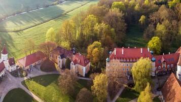 An aerial view of a historic estate with multiple buildings, including a large main house with a red roof, smaller structures, and a church with a tower.