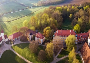 An aerial view of a historic estate with multiple buildings, including a large main house with a red roof, smaller structures, and a church with a tower.