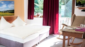 A bedroom with a bed, a nightstand, a chair, a table, a window with curtains, and a painting on the wall. - Kelten-Hotel (Unterbreizbach)