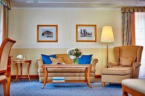 A hotel room with a sofa, armchair, small table, and a lamp. There are two framed pictures on the wall.