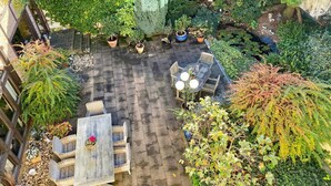 Un espace patio avec une table en bois et des chaises en osier, entouré de plantes en pot et d’un petit jardin.