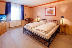 A double bed with a metal frame, a television on a wooden cabinet, a desk with a chair, and a lamp on a side table.