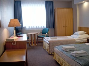 A hotel room with a bed, a desk with a lamp, a chair, a wardrobe, and a window with curtains.