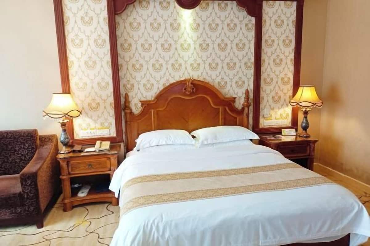 A hotel room with a large bed, two bedside tables with lamps, a wooden headboard, patterned wallpaper, and a brown armchair.