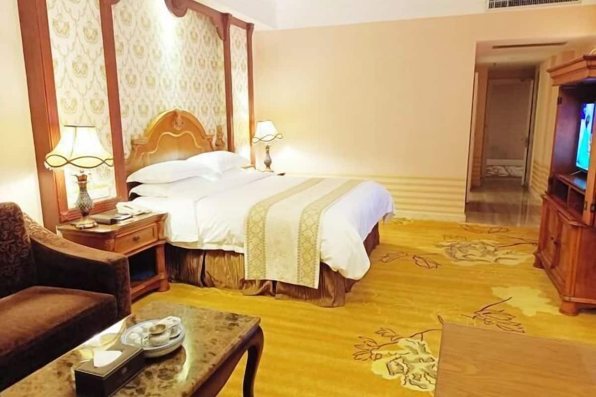 A hotel room with a bed, two bedside lamps, a television, a wooden nightstand, a chair, and a small table with a tray.