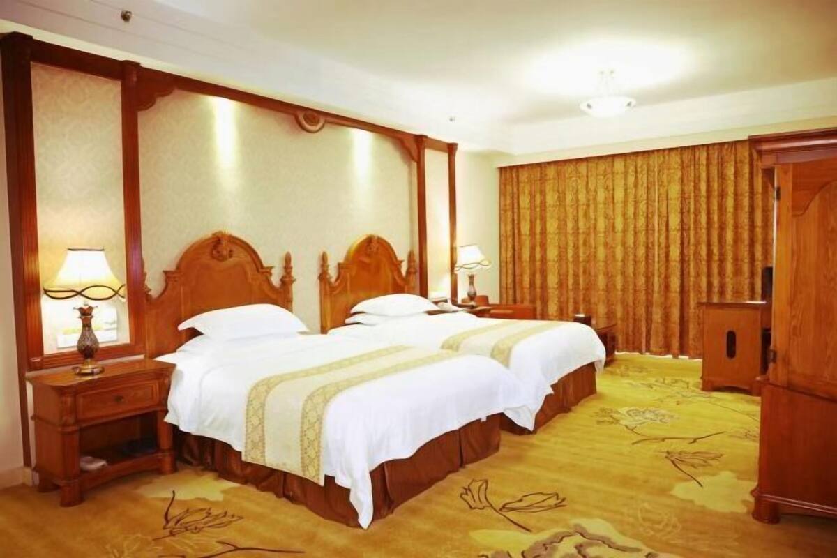 A hotel room with two beds, wooden headboards, a large mirror, a nightstand with a lamp, and a wardrobe.