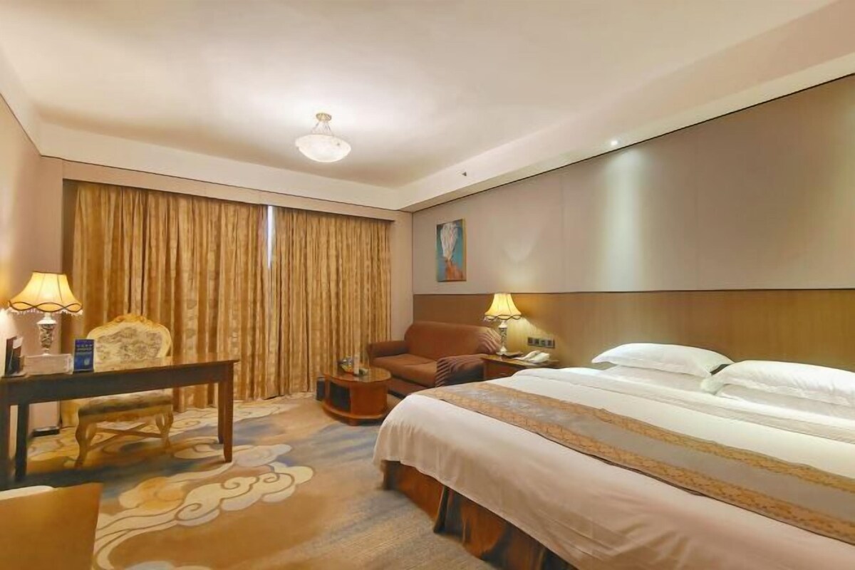 Una habitación de hotel con cama, escritorio, silla, lámpara y cortina.