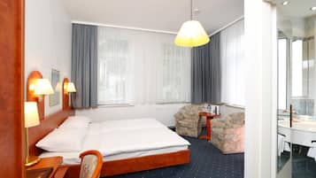 Ein Hotelzimmer mit Bett, Nachttischlampen, einem Sessel, einem kleinen Tisch, einem Spiegel und einem Fenster mit Vorhängen.