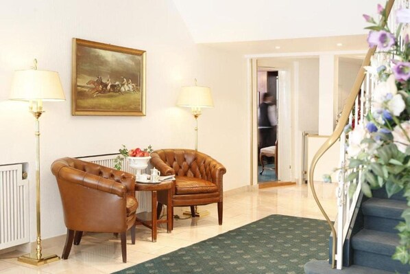 A room with a brown leather armchair, a small table with a teapot, two lamps, a framed painting, and a staircase with floral decorations. - Hotel Stadt Hannover (Goettingen)
