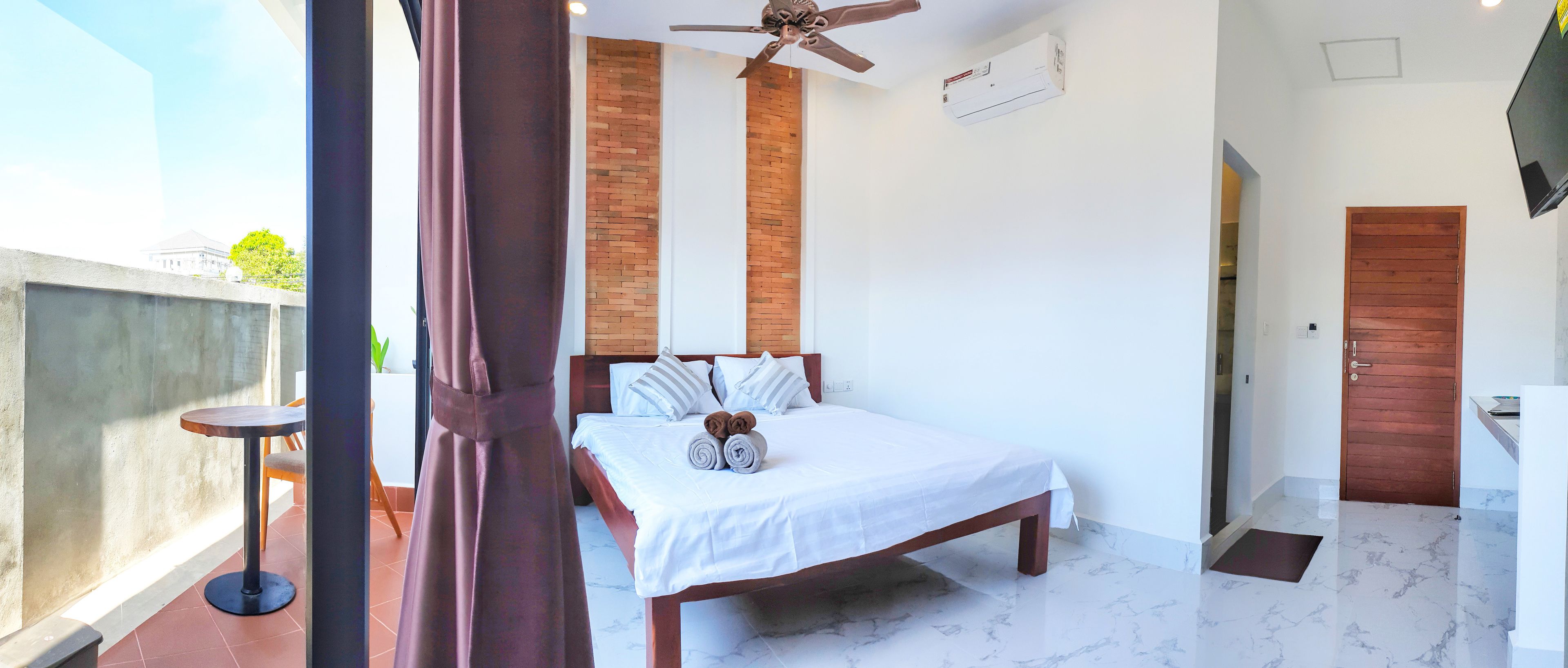 Un dormitorio con cama, ventilador de techo, pared de ladrillo, puerta de madera y baño con lavamanos.