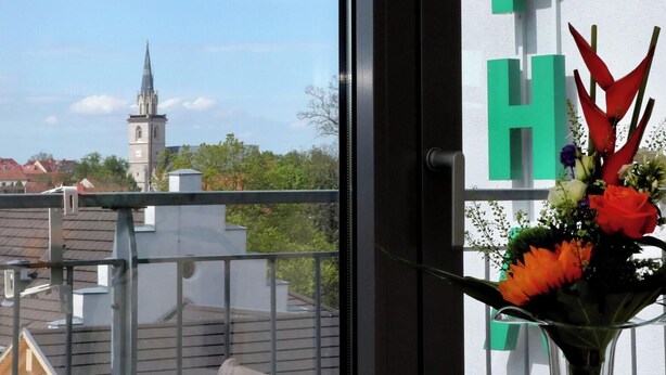 A balcony with a view of a church tower, a white wall with the letters 'ALPH' visible, and a vase with flowers. - Alpha (Bad Langensalza)