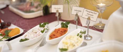 Una mesa servida con diversos platos etiquetados en alemán, incluyendo filete de hake, ensalada rusa casera y un plato con guarnición amarilla.