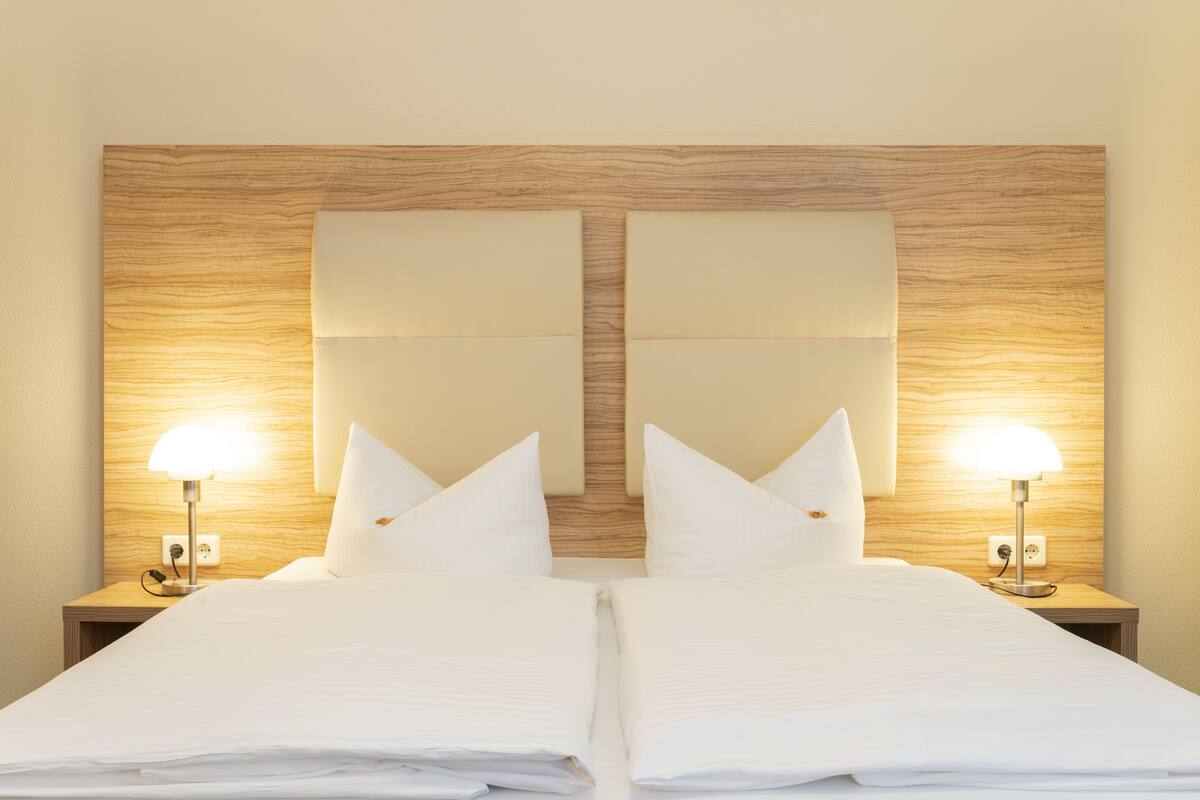 Una habitación de hotel con cabecera de madera, dos mesitas de noche con lámparas y una cama bien hecha con ropa blanca.