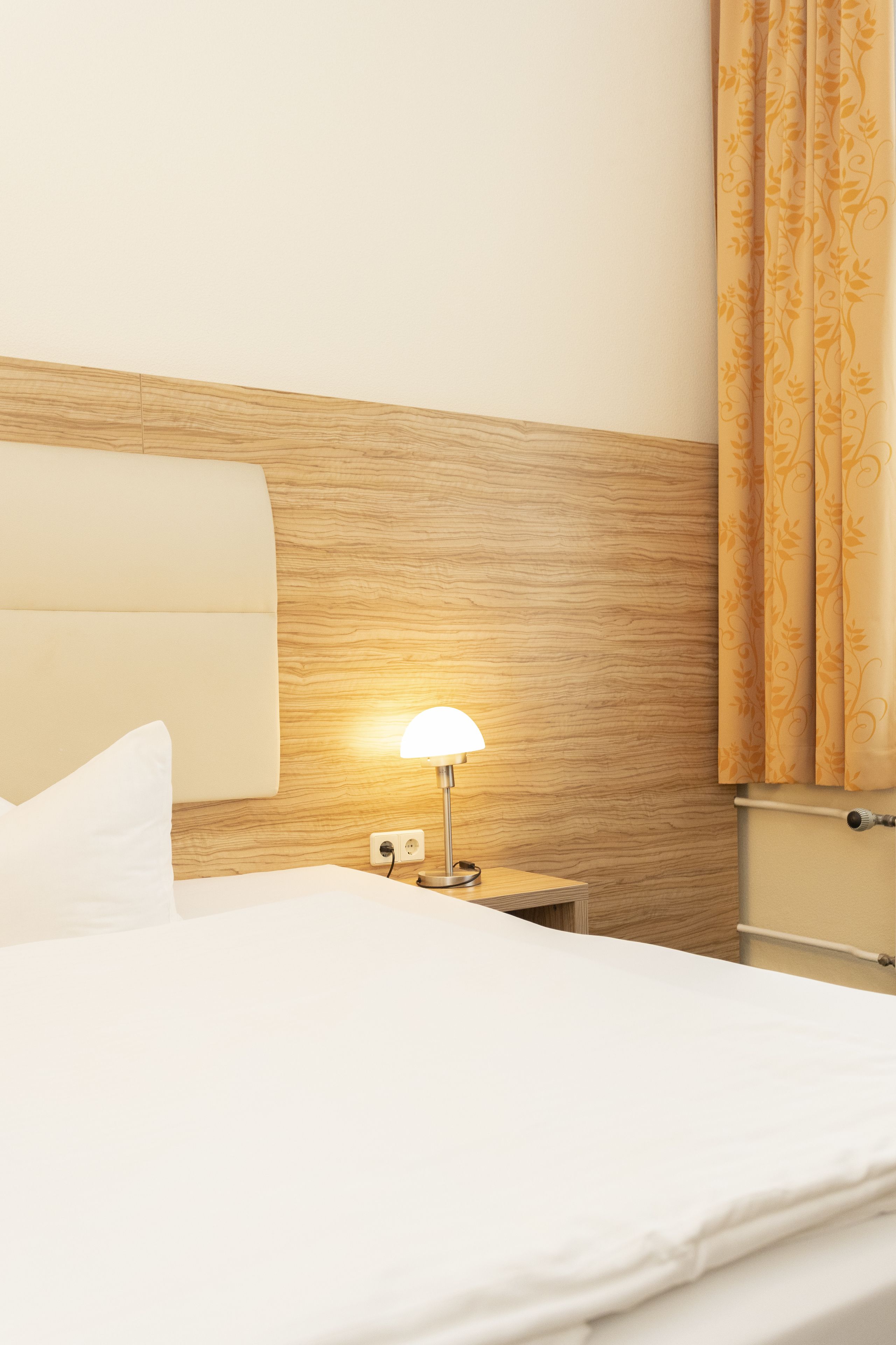 A neatly made bed with white linens, a wooden headboard, a bedside lamp, and a curtain with a floral pattern.