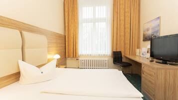 Quarto de hotel com cama, escrivaninha, cadeira, televisão e janela com cortinas.