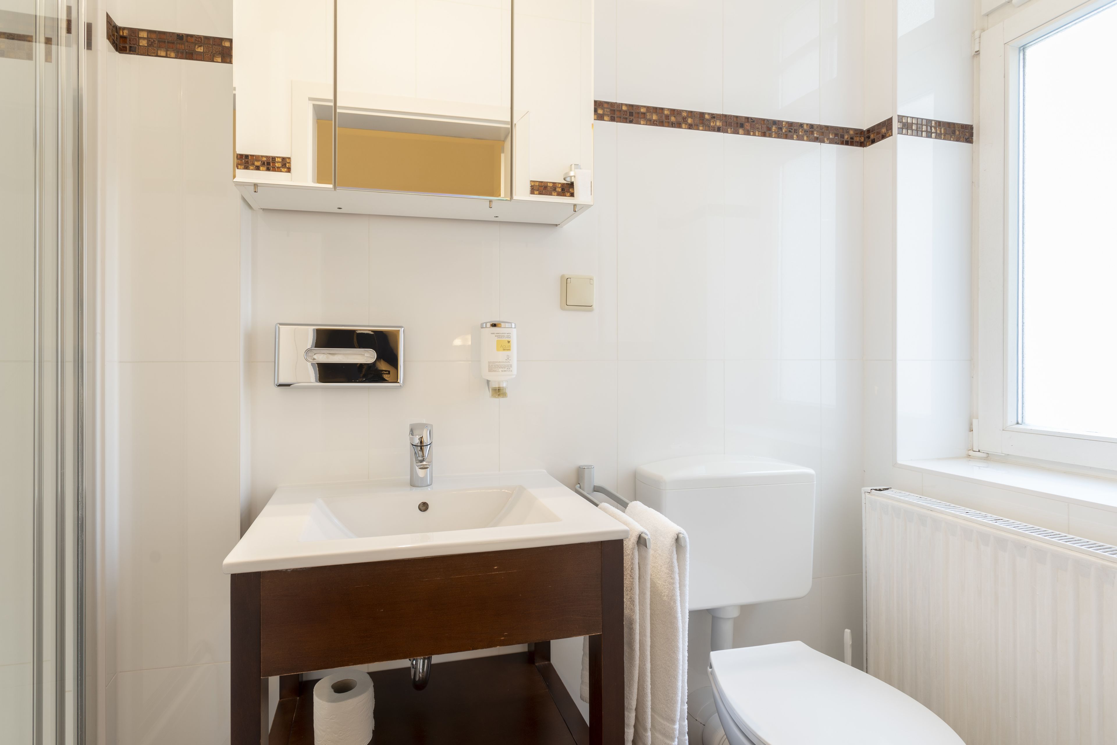 Un baño con lavamanos blanco, tocador de madera oscura, un espejo sobre el lavamanos, un inodoro y una ventana.