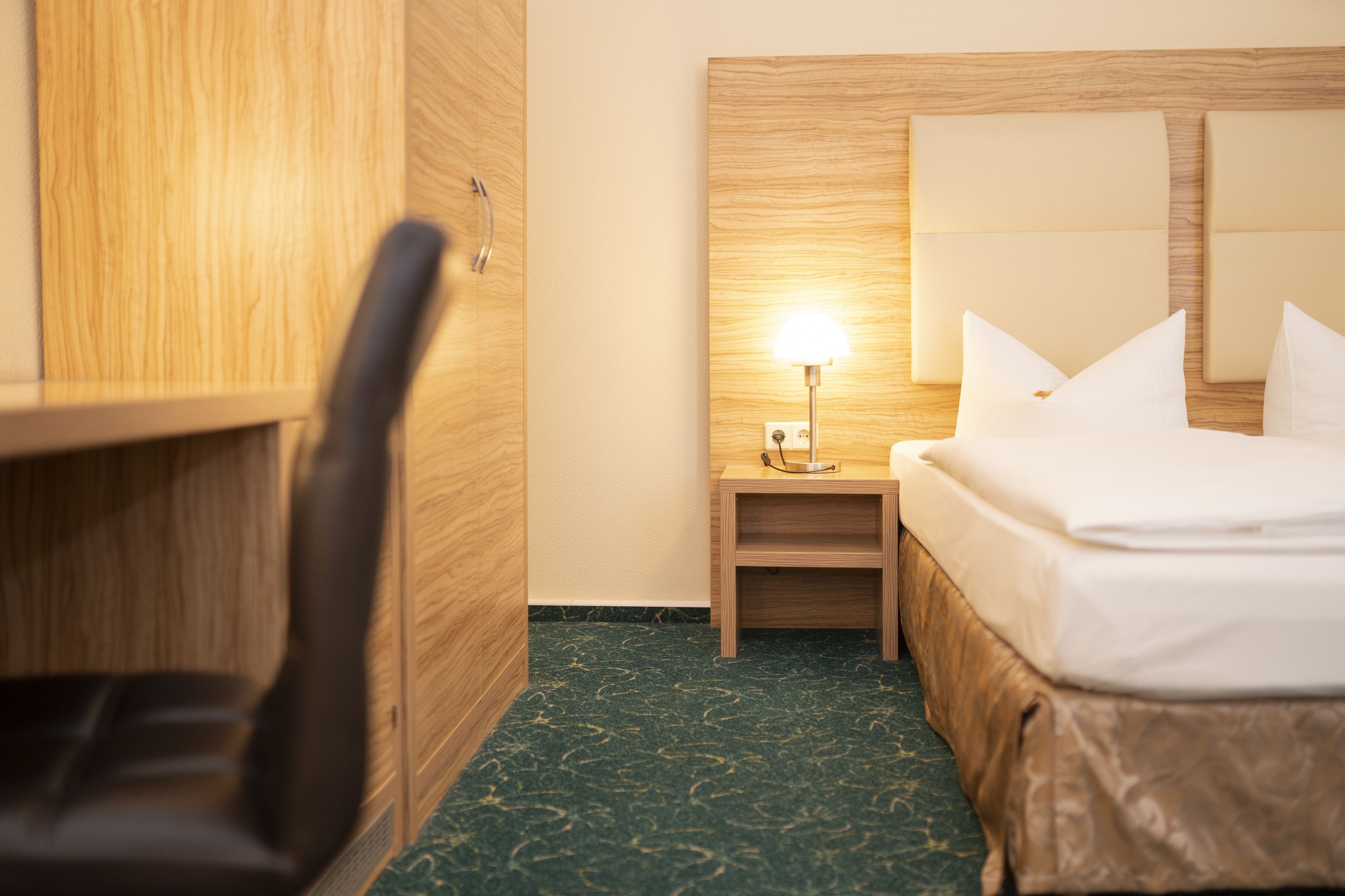 Une chambre d’hôtel avec une armoire en bois, une table de chevet avec une lampe, un lit avec du linge de maison blanc et une moquette verte.