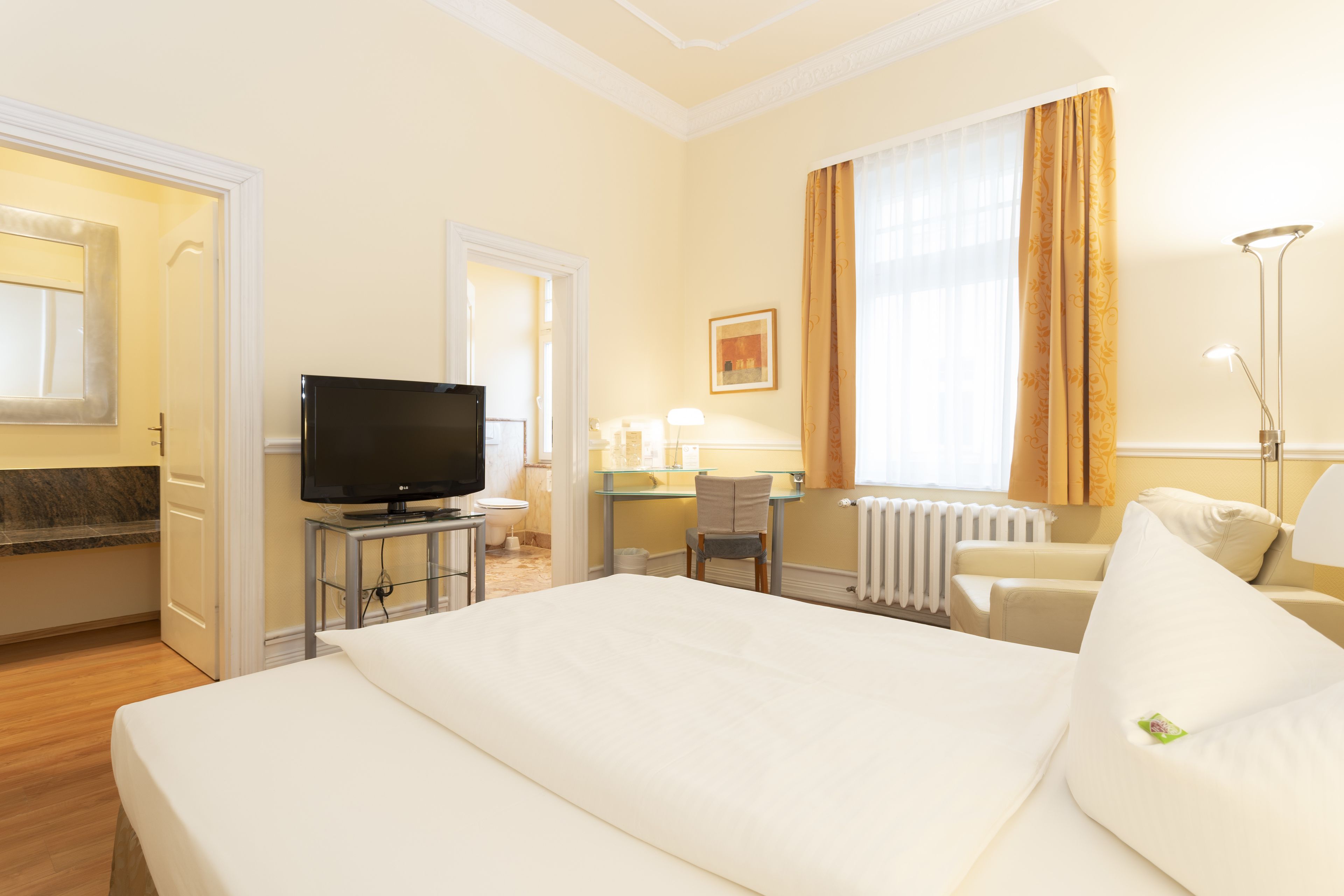 Habitación de hotel con una cama grande, televisión, escritorio con silla, ventana con cortinas y radiador.